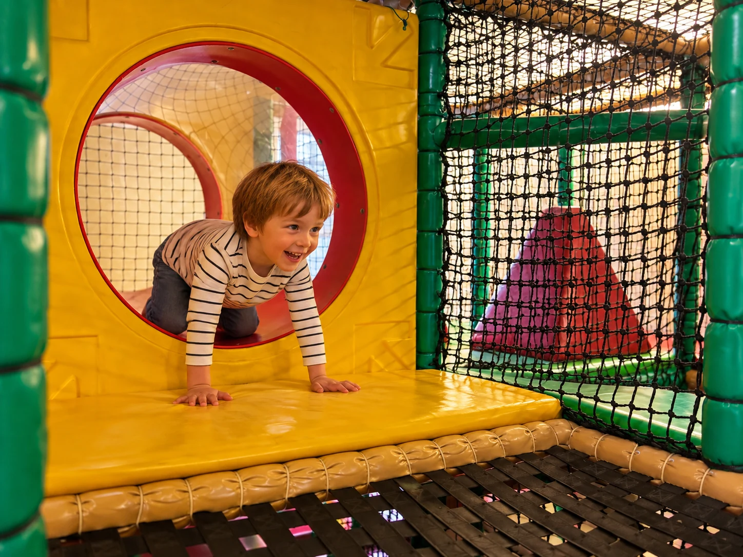 Lachender Junge krabbelt durch einen runden gelben Klettertunnel im Indoorspielplatz des Kindercafés Spielzimmer.